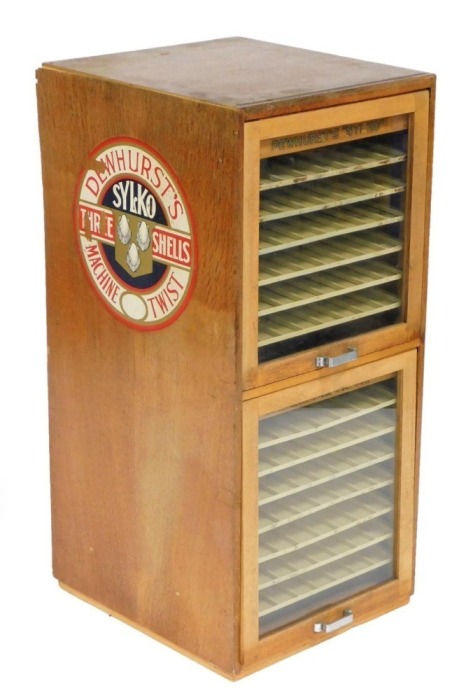 An early 20thC Dewhurst's Sylko Three Shells oak cased shop display cabinet, of two section form, each with an up and over glazed door enclosing six above seven shelves for cotton reels, 85cm high, 39cm wide, 43cm deep.
