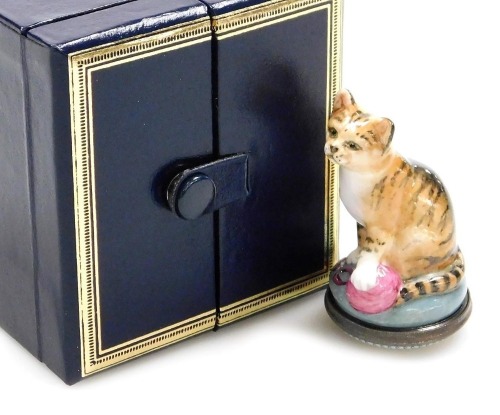 A Halcyon Days porcelain and enamel bonbonniere, modelled as a cat with one paw raised over a ball of wool, 5.5cm high, boxed with outer box.