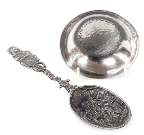 An early 20thC Keswick School of Industrial Arts hammered steel dish, with central incised decoration, stamped verso Keswick J976, 9cm diameter, together with a Dutch pewter spoon, the bowl with embossed decoration depicting figures, 20cm long.