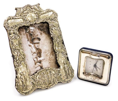 An early 20thC silver plated strut photograph frame, of military interest, embossed with soldiers, a battleship, an aeroplane, and armaments, etc., aperture 12.5cm x 8cm, frame 22cm x 16cm, together with a silver fronted bedside clock, stamped 925. (2)