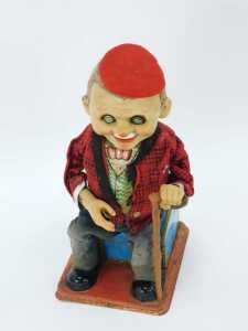 A 20thC tinplate figure of a gentleman seated with walking cane on Russian suitcase, by Roscoe, 30cm high. (AF) - 2