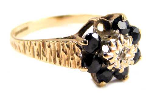 A 9ct gold sapphire and diamond cluster ring, set with seven sapphires and central tiny diamond, in a claw setting, on bark effect shoulders, one stone missing, ring size N&frac12;, 2.3g all in. (AF)