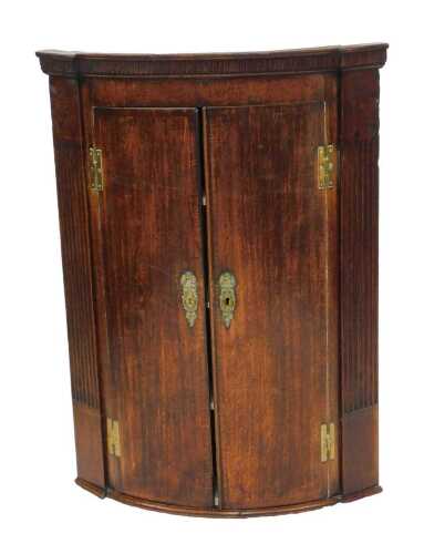 An early 19thC oak bow fronted corner cabinet, the top with a moulded edge above two doors with eagle surmounted brass escutcheons, flanked by reeded pilaster, 99cm high, 70cm wide, 35cm deep.