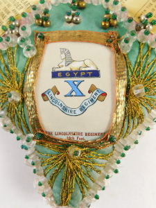 A Lincolnshire Regiment sweetheart heart shaped pin cushion, in pink silk with gold braid decoration, and regimental crest, printed ribbon text, 18cm. - 4