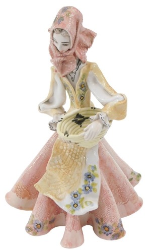 A modern French faience figure of a woman in a standing pose, holding a basket, in pink, ochre and blue, 23.5 cm high.