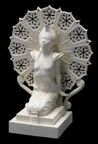 A cream glazed ceramic figure of a semi nude Oriental dancer, modelled kneeling on a plinth in...