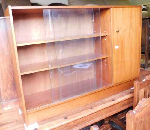 A teak bookcase, with two sliding glass doors flanked by a cupboard raised on out splayed legs....