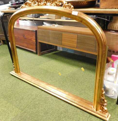 A Victorian-style arched gilt overmantel mirror with detailed corner scrolls, and a flat plinth...