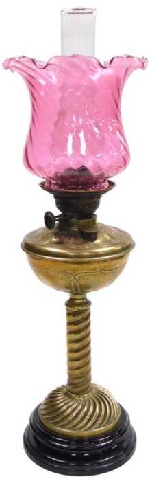 A Victorian brass oil lamp, the wrythen cranberry tinted shade, above a leaf decorated reservoir...