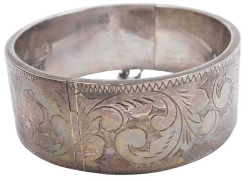 A silver bangle, with engraved foliate decoration, on a snap clasp, with safety chain as fitted,...