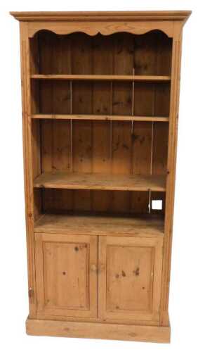 A pine cupboard bookcase, the outswept pediment over three shelves, above a pair of cupboard...