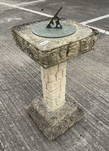 A stone sundial, with square top, metal central section, textured plinth and stepped base.