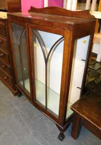An early 20thC oak display cabinet, the top with a shaped raised back, above arched glass doors...