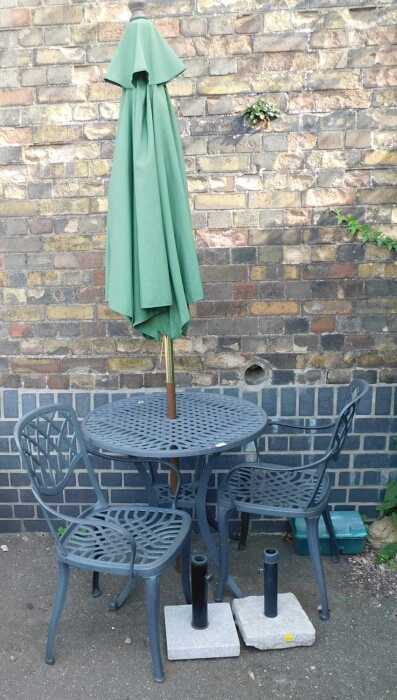 An aluminium garden bistro set, comprising a lattice work table, two chairs, and a parasol.