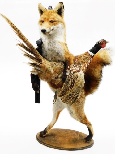 A taxidermy model of a fox, in hunting pose carrying pheasant with rifle, on an oval base, 95cm high.
