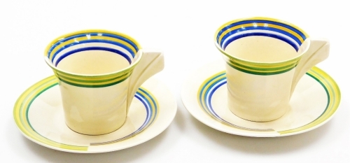 A pair of Clarice Cliff Bizarre coffee cans and saucers, of Art Deco design, each with a conical block handle and decorated with blue and yellow, and green and yellow repeat decoration, the saucer 12.5cm diameter, and the can 6cm high.