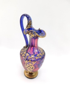 A late 19thC Bohemian pink and blue flashed glass ewer, decorated with shield reserves of deer and hunting dog, in a woodland setting, against a ground over decorated in gilt with trailing leaves, 33.5cm high. (AF) - 2
