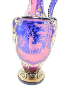 A late 19thC Bohemian pink and blue flashed glass ewer, decorated with shield reserves of deer and hunting dog, in a woodland setting, against a ground over decorated in gilt with trailing leaves, 33.5cm high. (AF) - 4