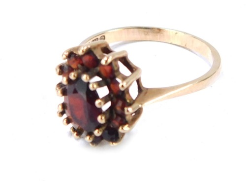 A 9ct gold garnet cluster ring, with large oval garnet surrounded by a row of smaller stones in a raised basket setting, size P, 2.4g all in.